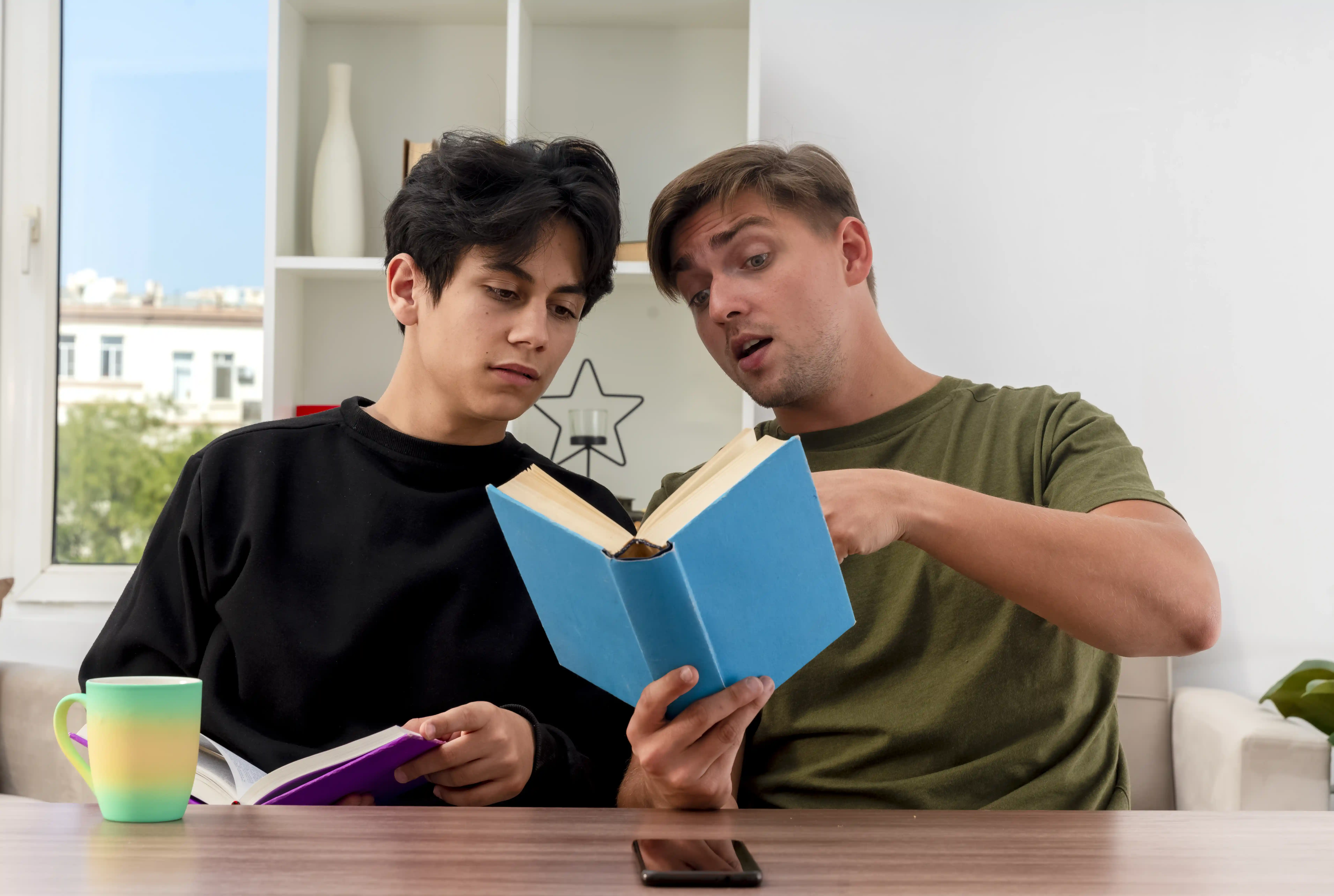 pleased-young-blonde-handsome-man-holds-points-book-sitting-table-with-young-brunette-handsome-boy-looking-book-inside-living-room.webp